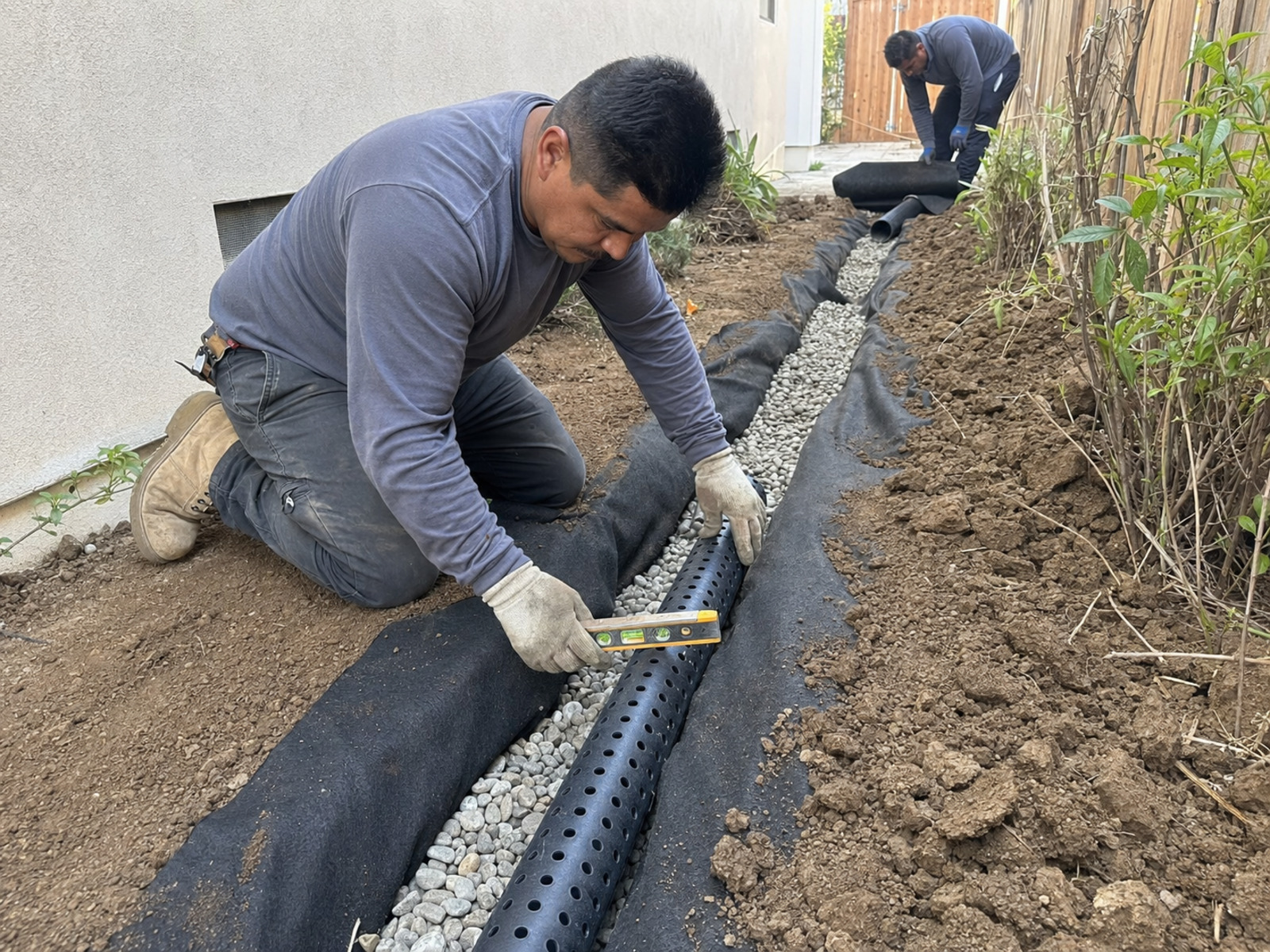 Worker leveling a gravel-filled drainage trench with a perforated pipe